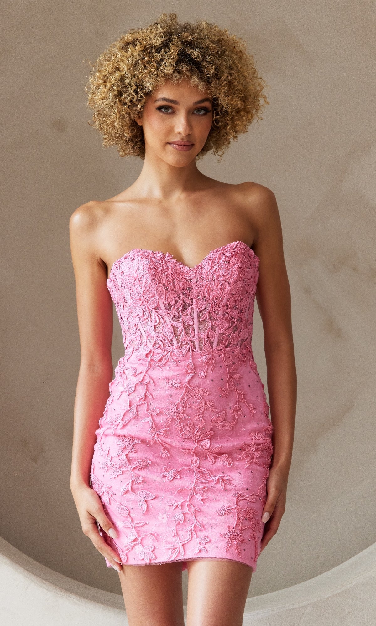 A woman with curly hair models the Embroidered-Lace Strapless Short Hoco Dress 7053S in pink, featuring a form-fitting sweetheart neckline, standing against a neutral background.